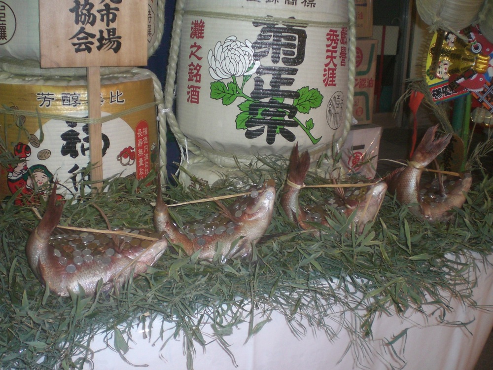 Des petits poissons morts recouverts de pièces de monnaie prennent la pose devant des bidons de sake (liqueur)...